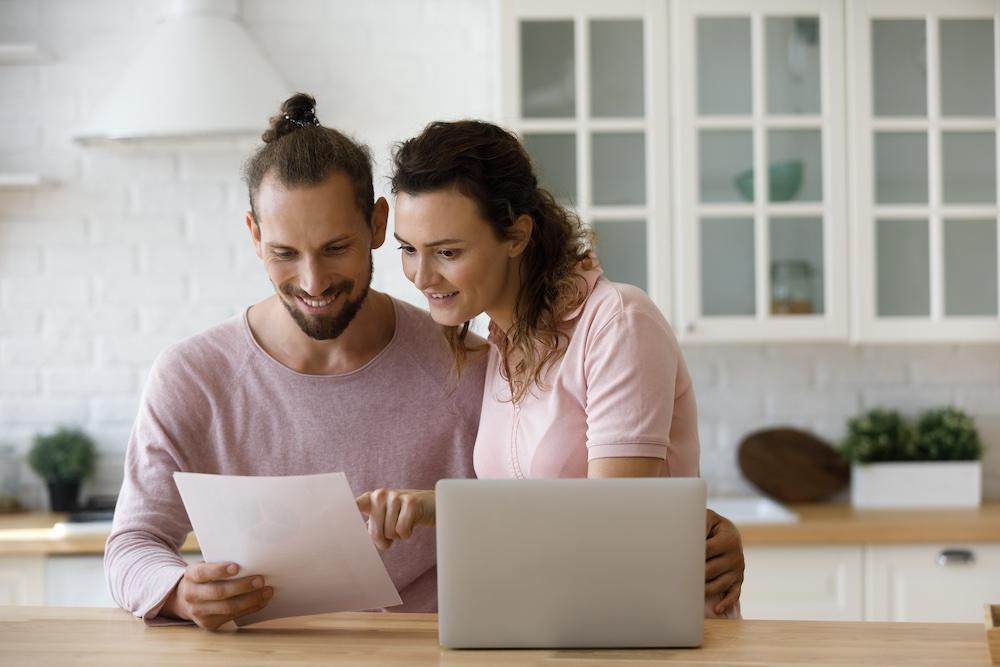Happy satisfied millennial married couple reading paper document at laptop in home kitchen, reviewing notice from bank, checking insurance, mortgage agreement with good terms